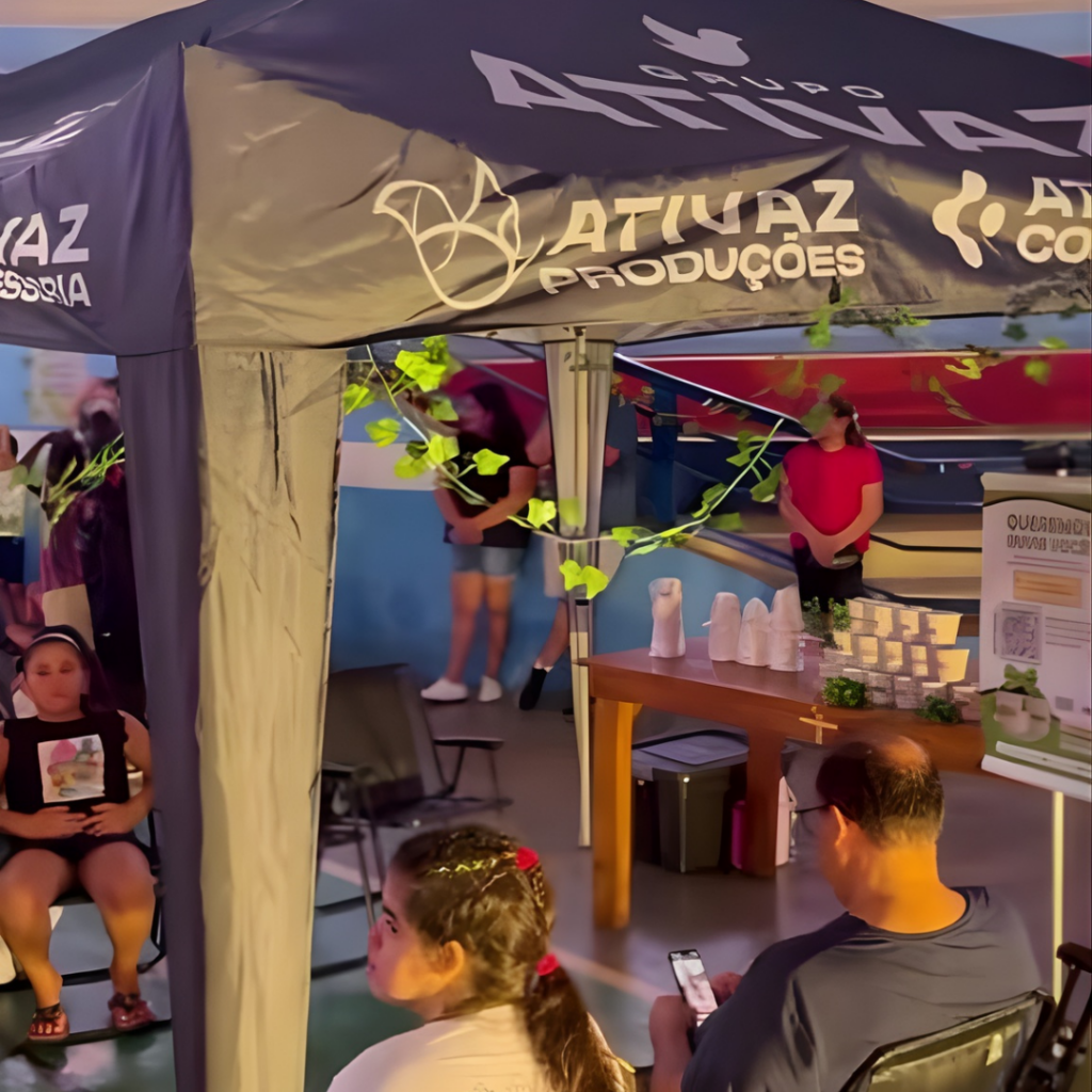 Ponto de apoio em evento cultural, com cadeiras e pessoas sentadas descansando, sob tenda institucional da Ativaz Produções, oferecendo acolhimento ao público durante a programação.