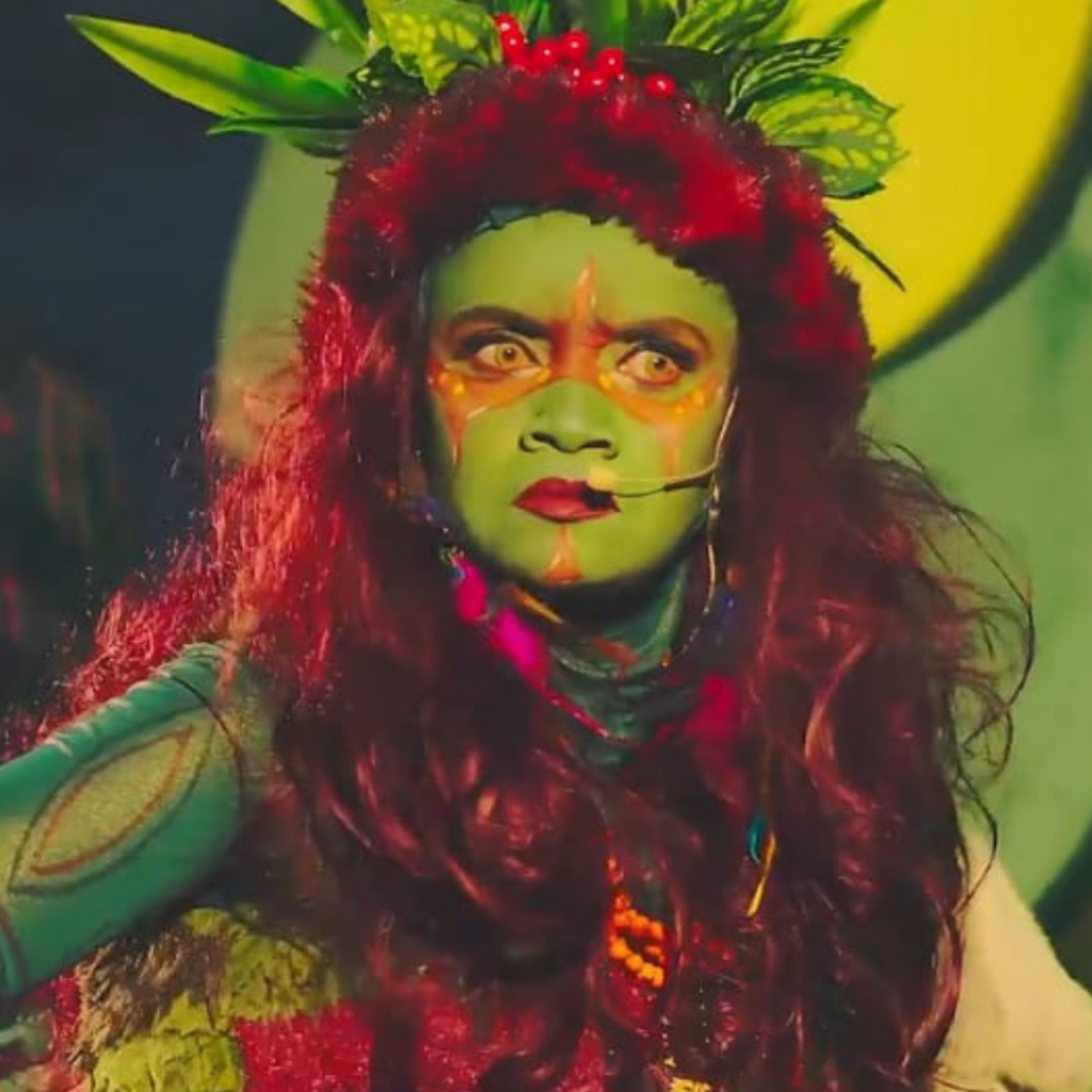 Atriz caracterizada como a personagem Caipora, do folclore brasileiro, com pintura corporal em tons de verde e vermelho, adereços de folhas e sementes na cabeça e figurino inspirado na fauna e flora da mata. A intérprete utiliza microfone de cabeça e atua em cena sob iluminação cênica colorida, com expressão intensa e foco dramático.