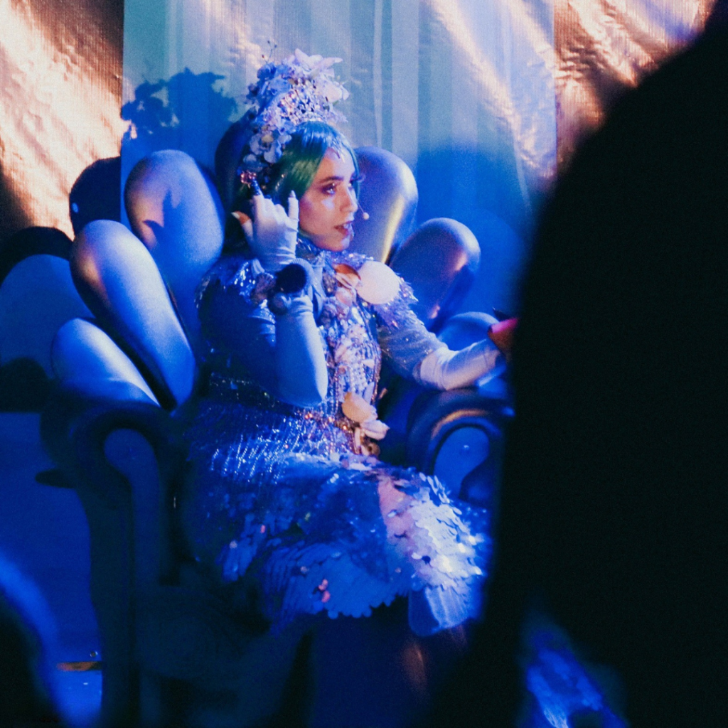 Atriz caracterizada como a personagem Iara, do folclore brasileiro, sentada em um trono cenográfico com formas orgânicas que remetem a conchas e ao fundo do rio. A intérprete utiliza figurino azul cintilante com aplicações brilhantes, adereços inspirados em elementos aquáticos e maquiagem temática. A cena ocorre sob iluminação azul, criando atmosfera aquática e mágica durante a apresentação teatral.