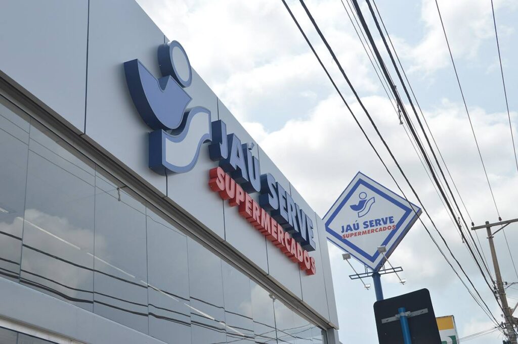 Fachada do supermercado Jaú Serve, com o logotipo em destaque na parede e a placa elevada da marca visível ao fundo, sob um céu parcialmente nublado.