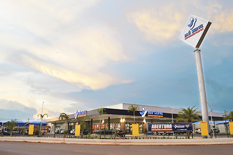 Fachada de uma unidade do supermercado Jaú Serve, com prédio amplo e moderno, estacionamento em frente, palmeiras ao redor e totens com a logomarca. Há faixas indicando abertura da loja. O céu está parcialmente nublado, com iluminação natural no ambiente.