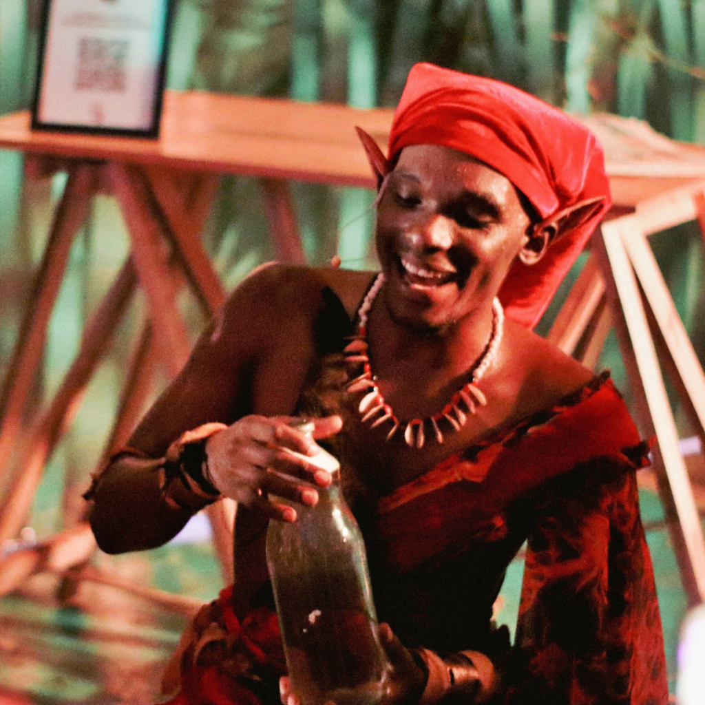 Ator caracterizado como Sací, personagem do folclore brasileiro, utilizando figurino em tons de vermelho, colar artesanal e adereços cênicos, durante apresentação teatral em espaço cenográfico com iluminação cênica. O intérprete segura um recipiente de vidro, interagindo com o público em cena.