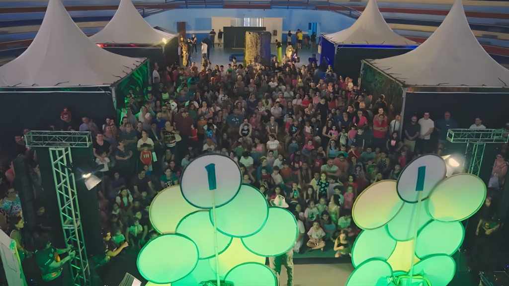 Vista aérea do público reunido no circuito cultura e familia, em um ginásio com estruturas cenográficas iluminadas em verde e tendas brancas ao fundo.