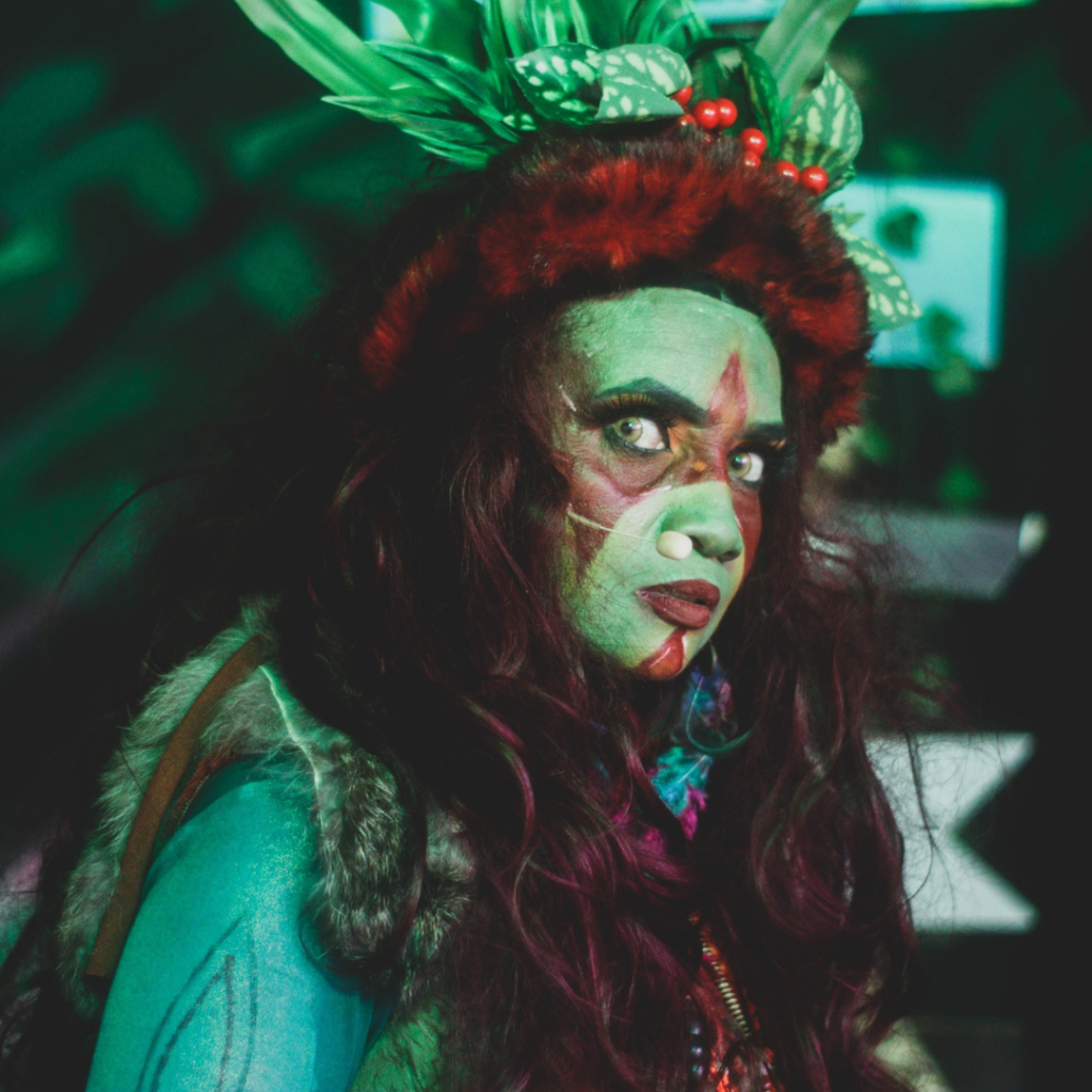 Atriz caracterizada como a personagem Caipora, do folclore brasileiro, com pintura corporal em tons de verde e vermelho, adereços de folhas e sementes na cabeça e figurino inspirado na fauna e flora da mata. A intérprete utiliza microfone de cabeça e atua em cena sob iluminação cênica colorida, com expressão intensa e foco dramático.