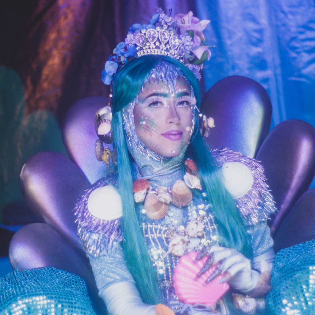 Atriz caracterizada como a personagem Iara, do folclore brasileiro, sentada em um trono cenográfico com formas orgânicas que remetem a conchas e ao fundo do rio. A intérprete utiliza figurino azul cintilante com aplicações brilhantes, adereços inspirados em elementos aquáticos e maquiagem temática. A cena ocorre sob iluminação azul, criando atmosfera aquática e mágica durante a apresentação teatral.