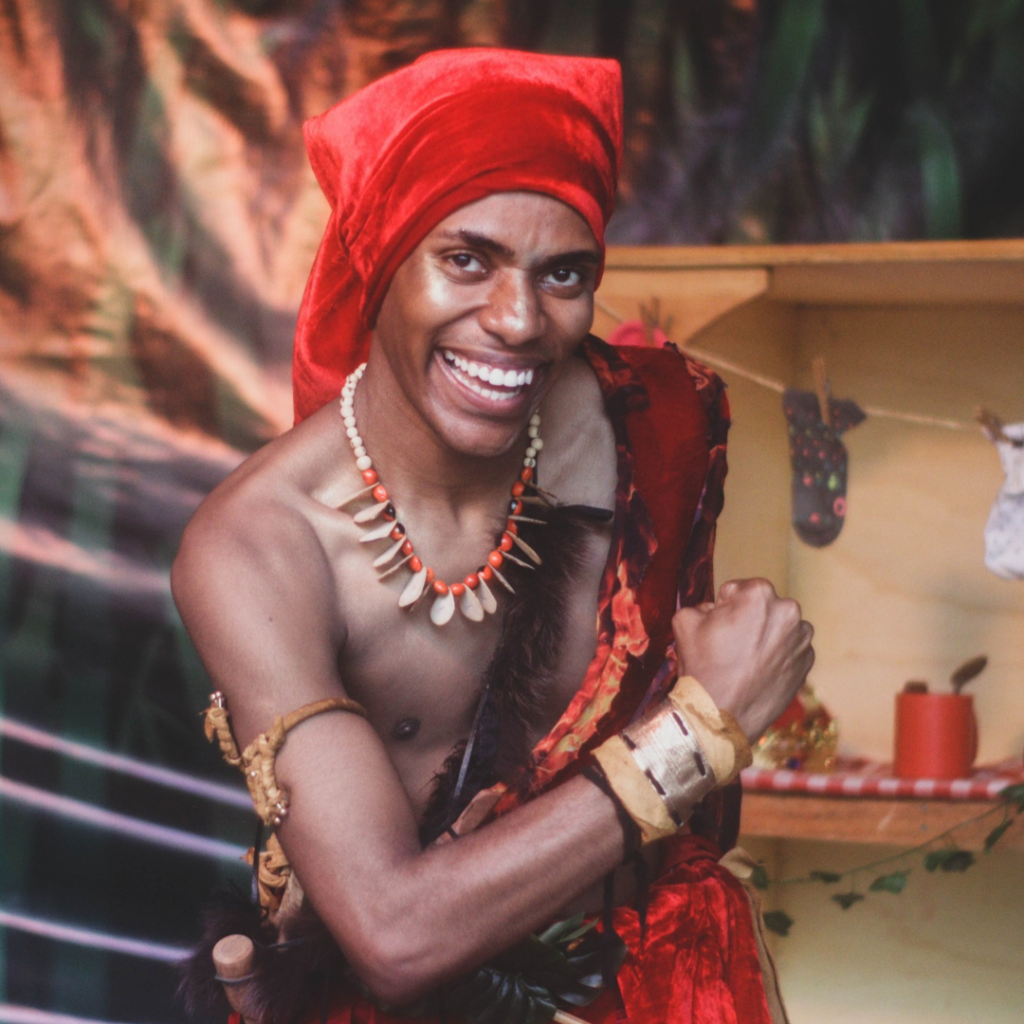 Ator caracterizado como Sací, personagem do folclore brasileiro, utilizando figurino em tons de vermelho, colar artesanal e adereços cênicos, durante apresentação teatral em espaço cenográfico com iluminação cênica. O intérprete segura um recipiente de vidro, interagindo com o público em cena.